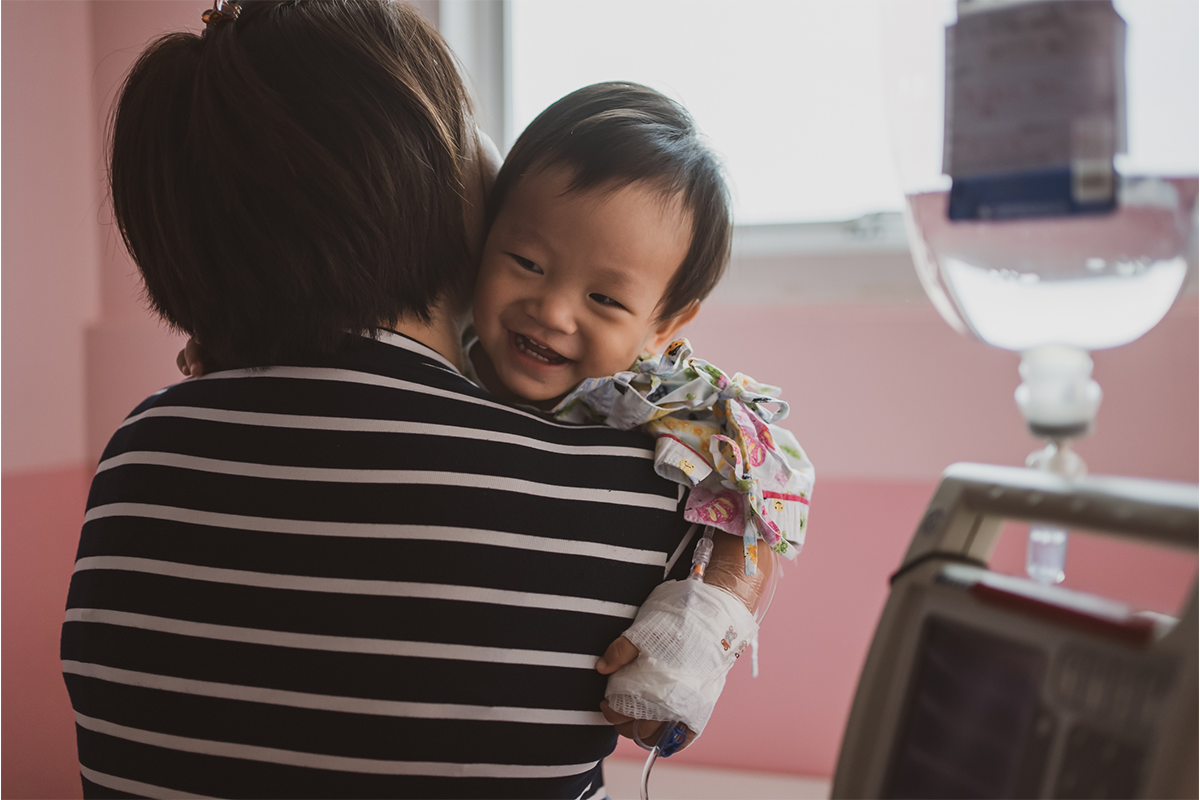 An adult holds a small child with a bandaged hand and an IV line in a hospital room.