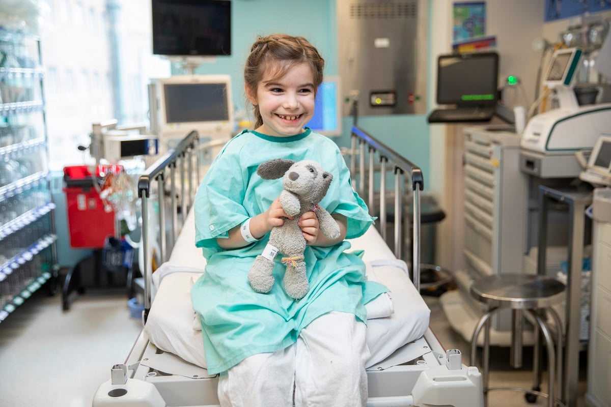 A child in a hospital gown sits on a bed holding a stuffed animal in a medical room.