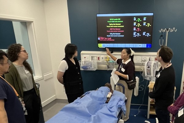 Healthcare staff stand around a patient on a hospital bed during a training session. A nurse points to a screen showing patient care information.