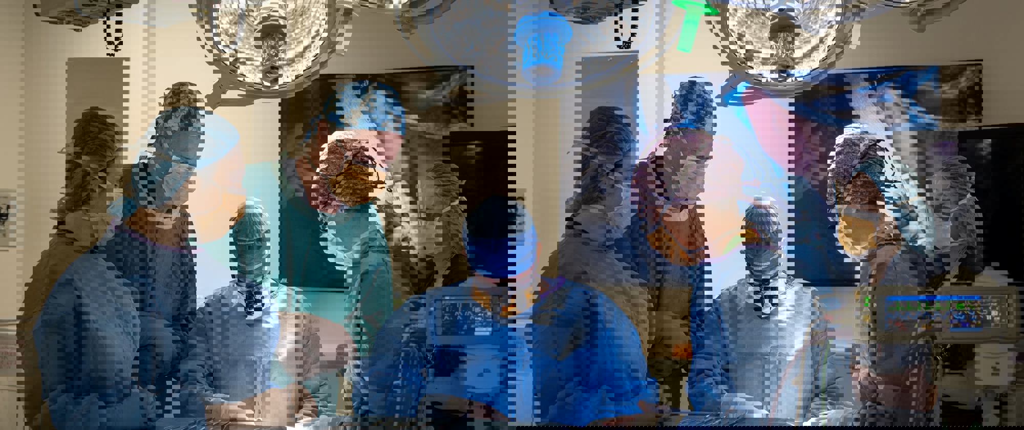 4 surgeons and assistants at the operating table with a digital display in the background and a bright light overhead