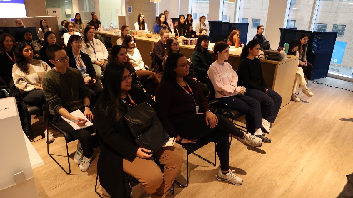 Audience seated in a well-lit room with large windows, attentively facing forward during an educational or professional event.