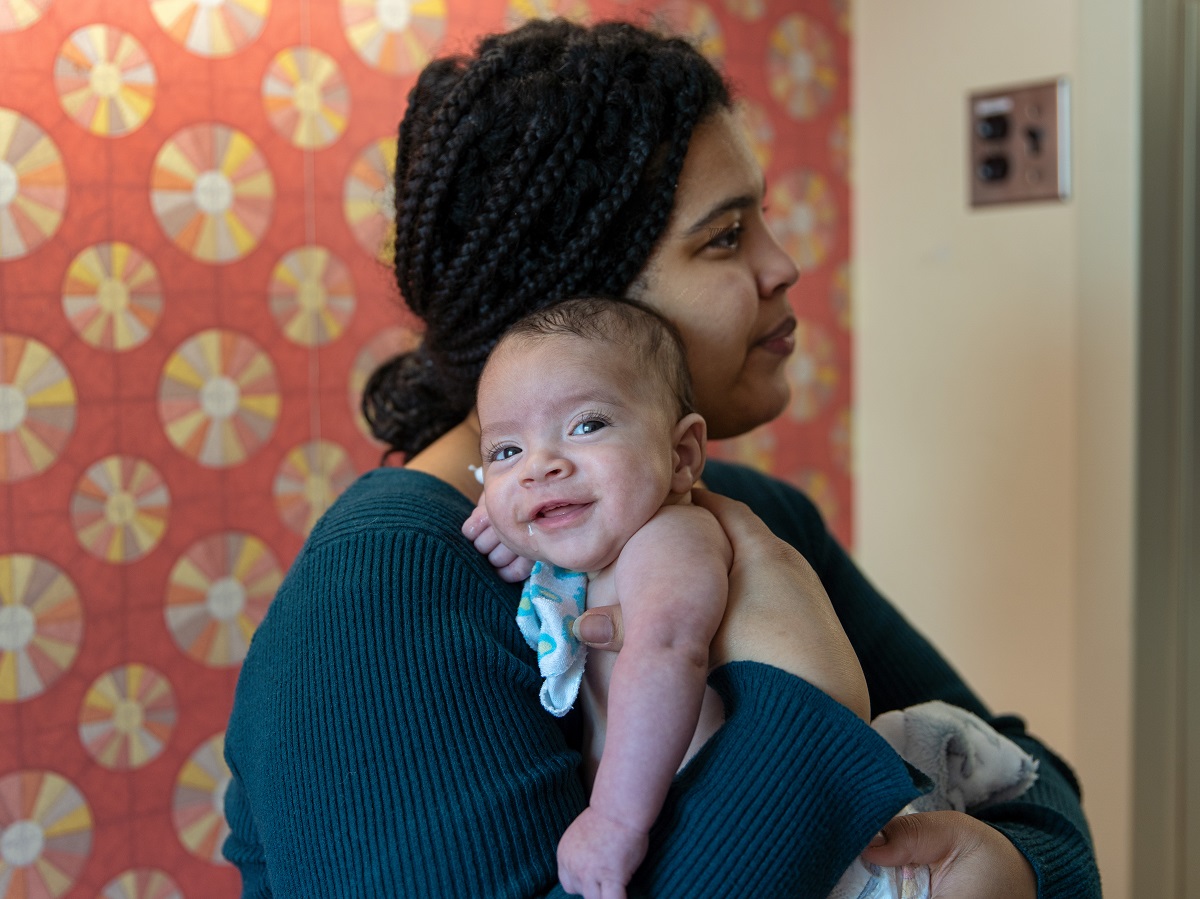 An adult holds a small baby against their shoulder in a room with patterned wallpaper.