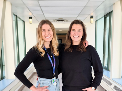 Two smiling individuals stand side by side in a sunlit hallway with large windows.