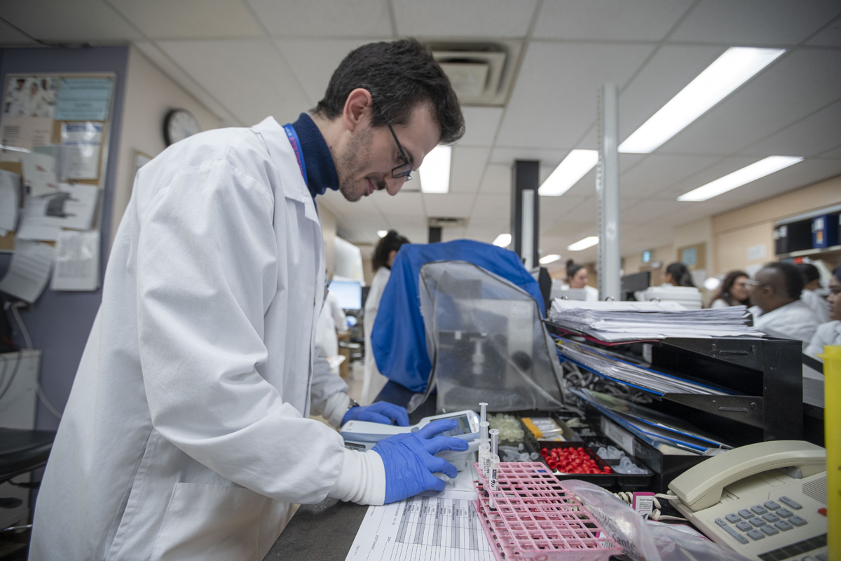 A SickKids staff member wearing a lab coat and blue gloves is working at a lab bench. In the background, other lab personnel are engaged in their tasks.