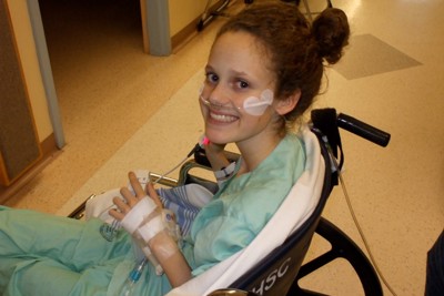 Young teenage girl with a bandage on her face smiling while seated in a wheelchair, wearing a hospital gown with medical tubes visible around her neck area.