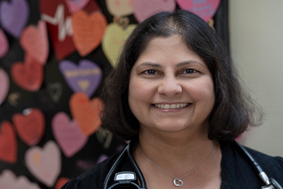 Wearing a black shirt and stethoscope, Anne smiles in front of colourful paper hearts on the wall.
