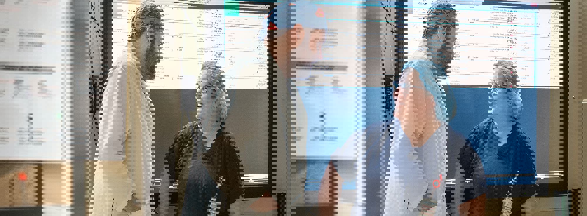 Two SK staff in PPE talking in a hallway in front of a digital chart display