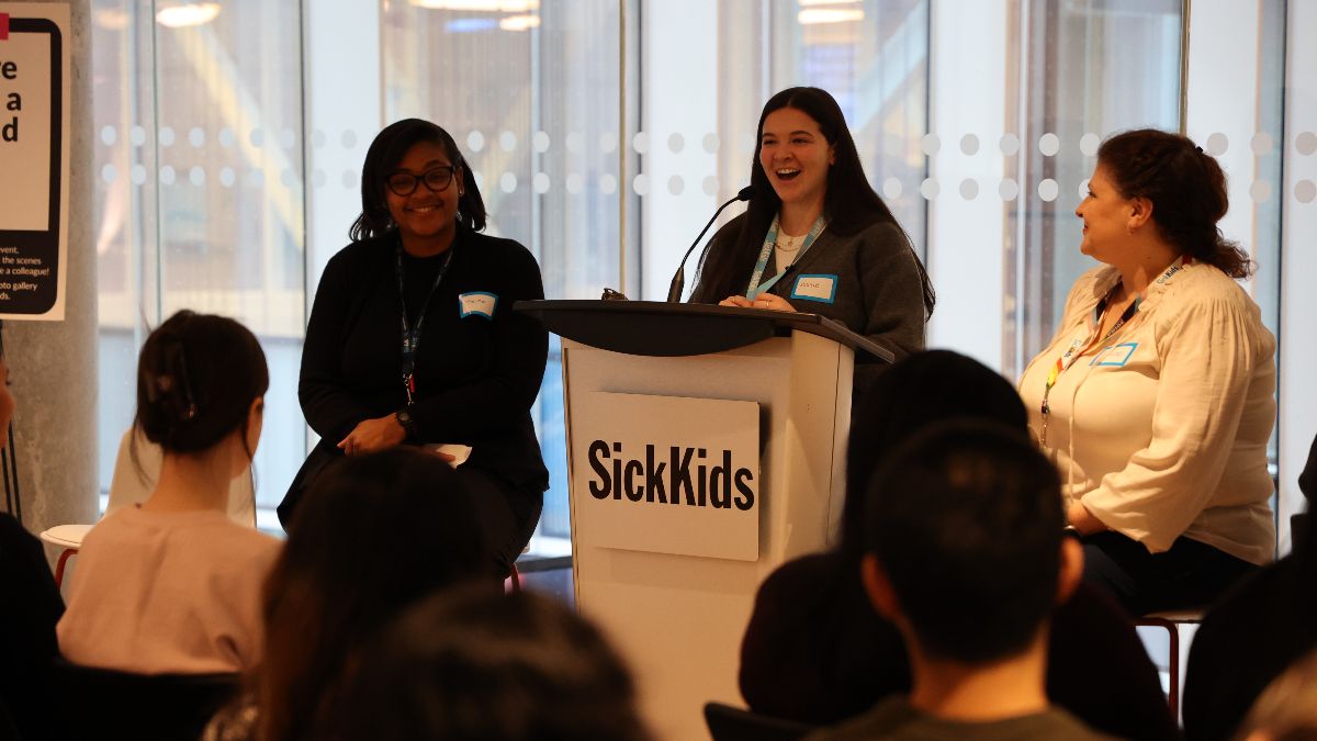Three speakers on a stage at SickKids, with one speaking at a podium and two seated beside, all smiling.