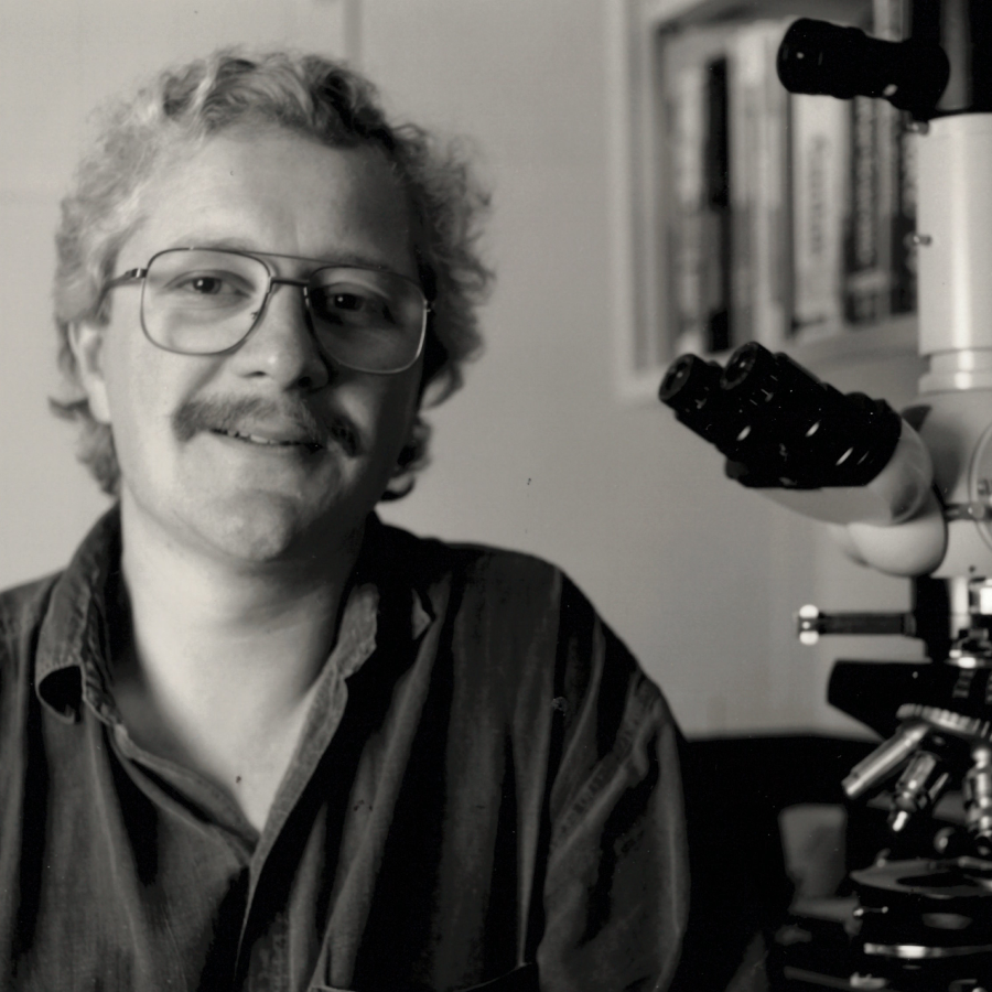 Black and white portrait of a smiling scientist with curly hair and glasses next to a microscope. He appears to be in a research laboratory setting.