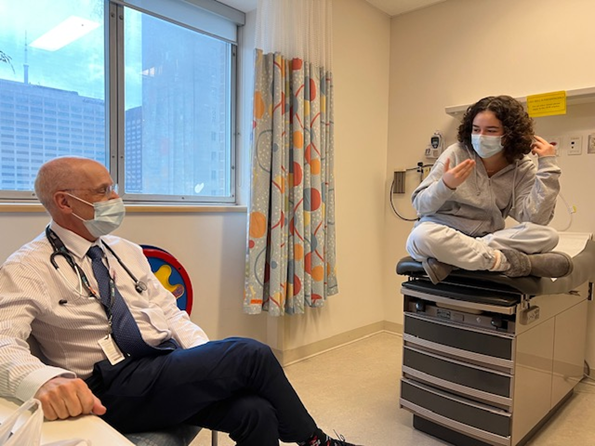 Color photo of a doctor wearing a mask consulting with a young patient in a hospital room. The patient sits on an exam table with city buildings visible through window.