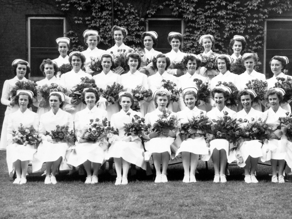  Graduation or capping ceremony photograph showing three rows of nursing students in white uniforms with caps, all holding floral bouquets in front of an ivy-covered building.