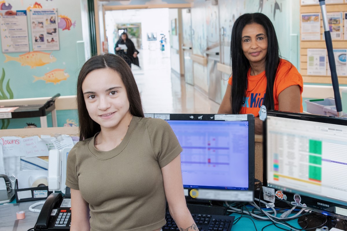 Two people stand on either side of a desk with counter and computers.