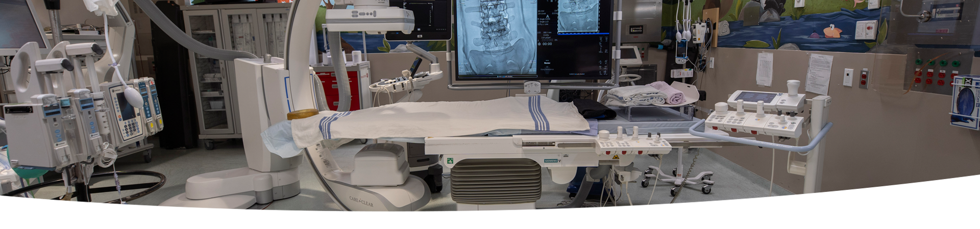 A room with a Single Plane Digital Angiography unit. There is a variety of hospital equipment in the background.