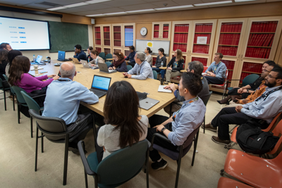 A group of about 20 people sitting around a table in a boardroom. Many of them have their laptops open or looking toward a large screen at the front of the room that is displaying a presentation slide.