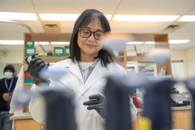 A lab professional wearing a lab coat and black gloves using a large pipette.