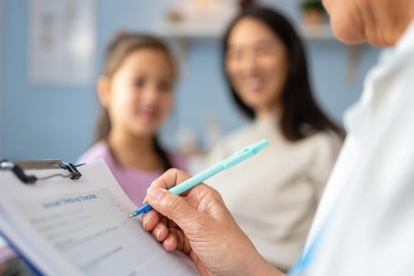 A healthcare professional holds a clipboard and pen while reviewing a form, with two patients seated nearby in a clinical exam room