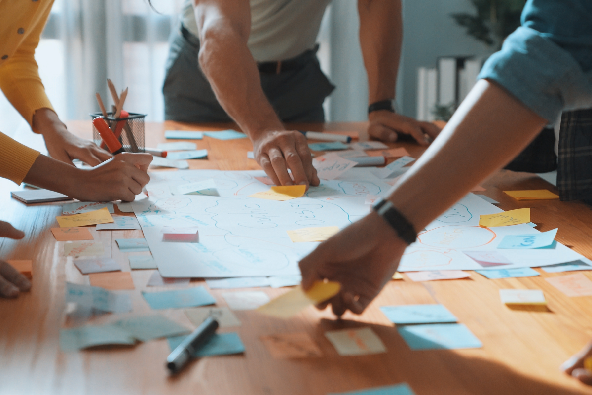 Many hands contributing to a paper on a table with markers and sticky notes.