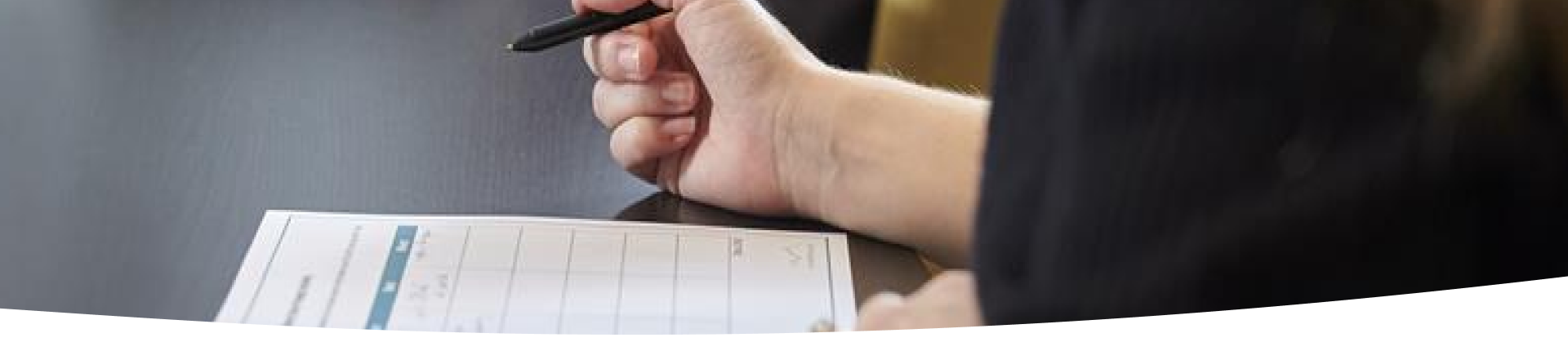 A person wearing a dark long-sleeve shirt is writing with a pen on a grid-layout form or worksheet placed on a dark table. Only the hand and part of the arm are visible.