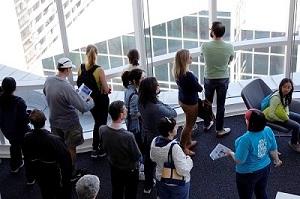 A group of adults look out through a floor to ceiling window in a tall building