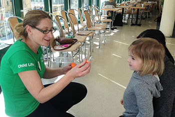 A woman wearing a Child Life tshirt kneels and holds a bubble wand while a child smiles standing next to her