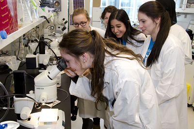 In a lab, a group of teenage girls wearing lab coats observe as another teen looks through a microscope