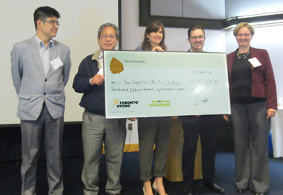 A group of five adults stand together smiling while holding a large cheque addressed to SickKids