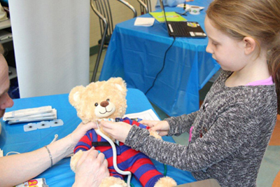 A young girl holds a medical thermometer against a teddy bear