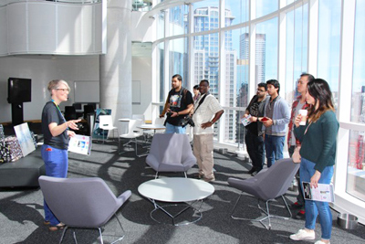 People stand in a brightly lit space with windows. One person speaks to the group.