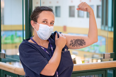 Karen Milford wearing scrubs and a face mask, flexes her arm to reveal an artistic bicep tattoo of a brain and flowers