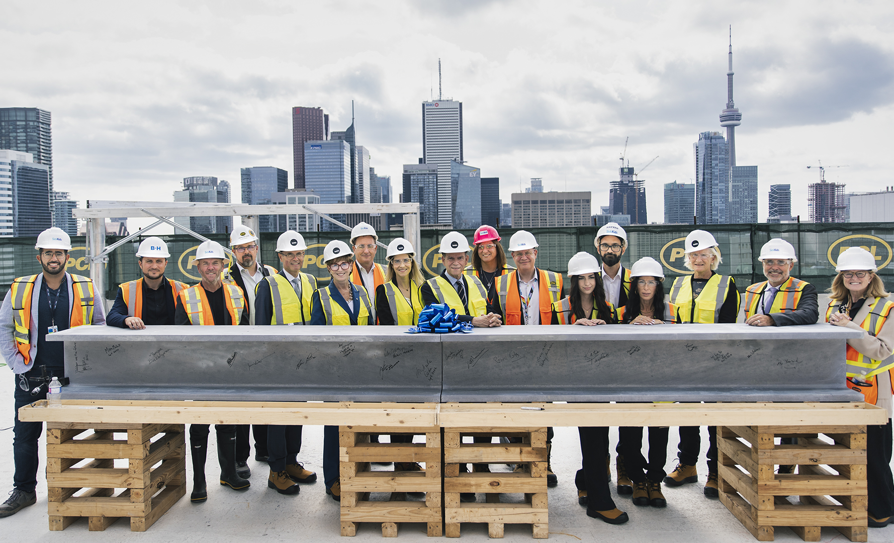 A large group of men and women wearing construction gear stand together with a large steel beam covered in signatures. The Toronto skyline is visible behind them