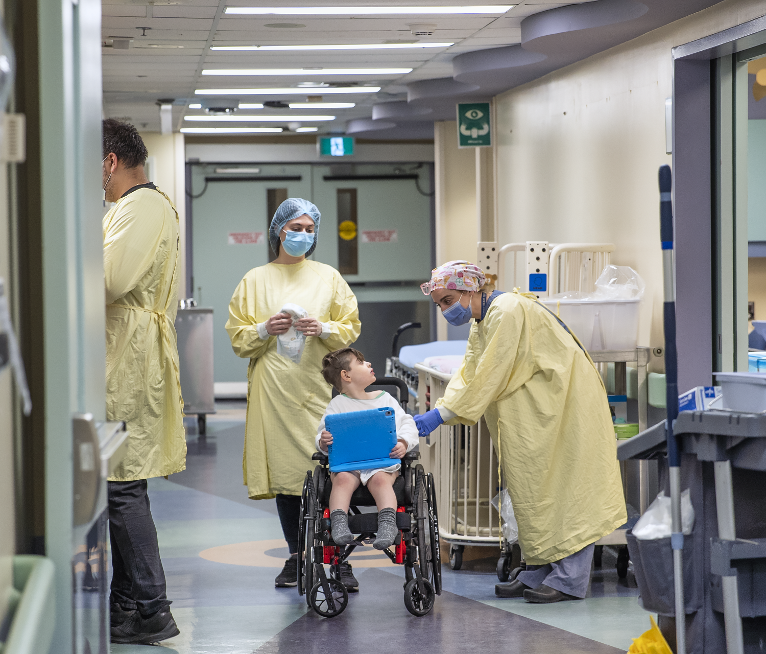 Child sits in a wheelchair holding a tablet. Three adults in medical gowns, caps and masks are present. One leans over and speaks to the child.