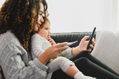 A person sits on the couch with a baby on their lap, holding and looking at their phone in their hand.