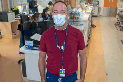 Social worker posing for the camera in front of the nursing station, hands in pockets, with Emergency Department in the background.