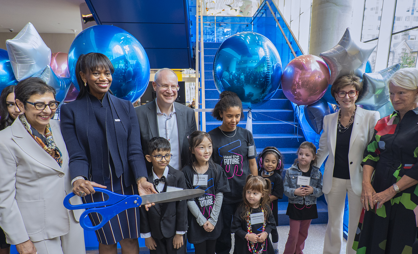 A group of children and adults, including one adult holding a large pair of scissors, gathered around a staircase that is roped off with a large ribbon and balloons