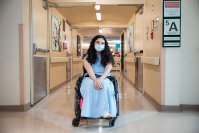Melika sitting in a wheelchair in the middle of a hospital hallway. A sign on the wall says "5A".