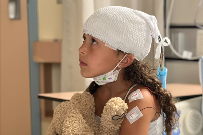 A girl sitting on a hospital bed while holding a teddy bear on her lap.