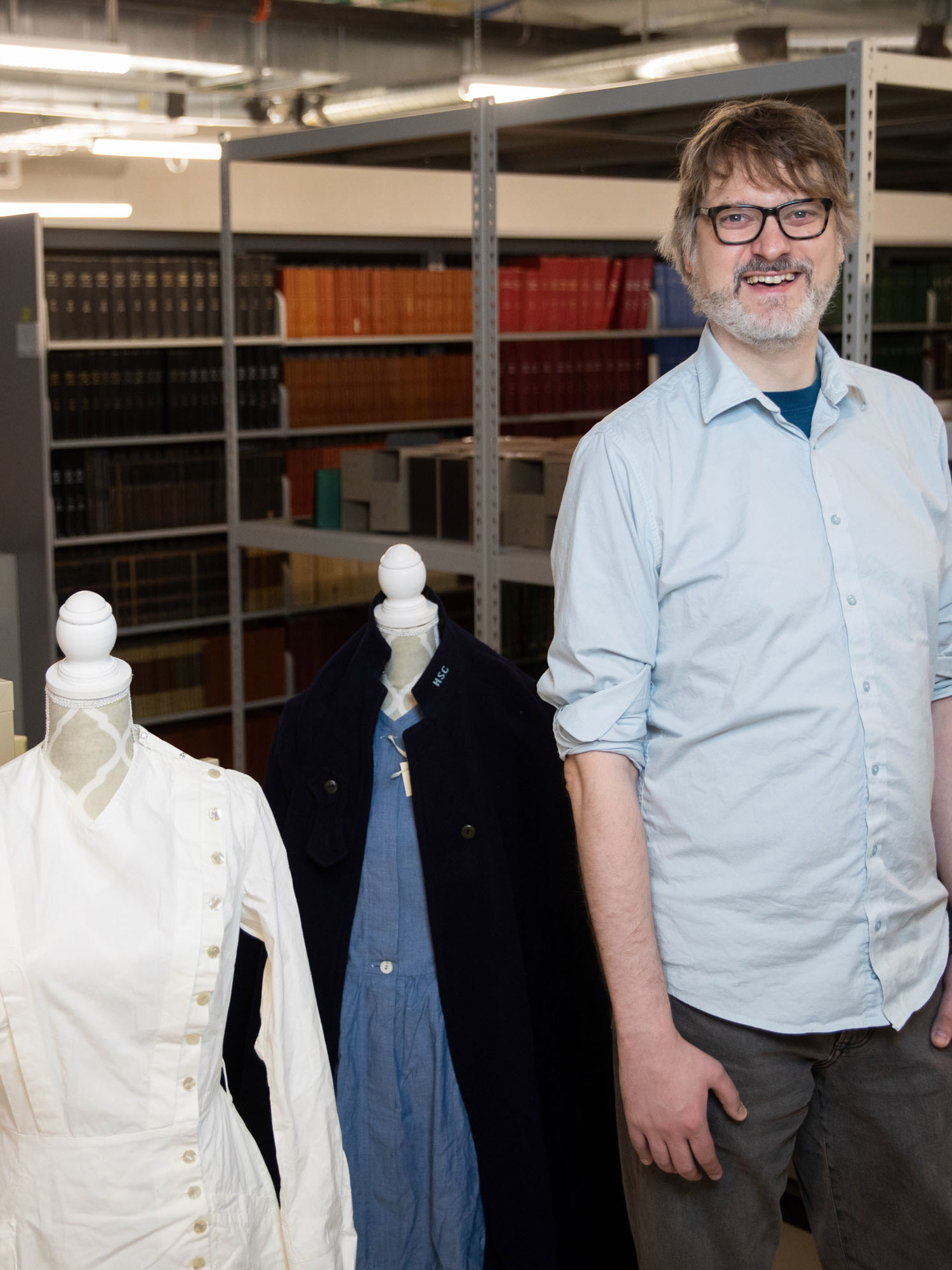 Man with glasses and light blue shirt standing in archive storage area with historical clothing displayed on mannequins nearby.