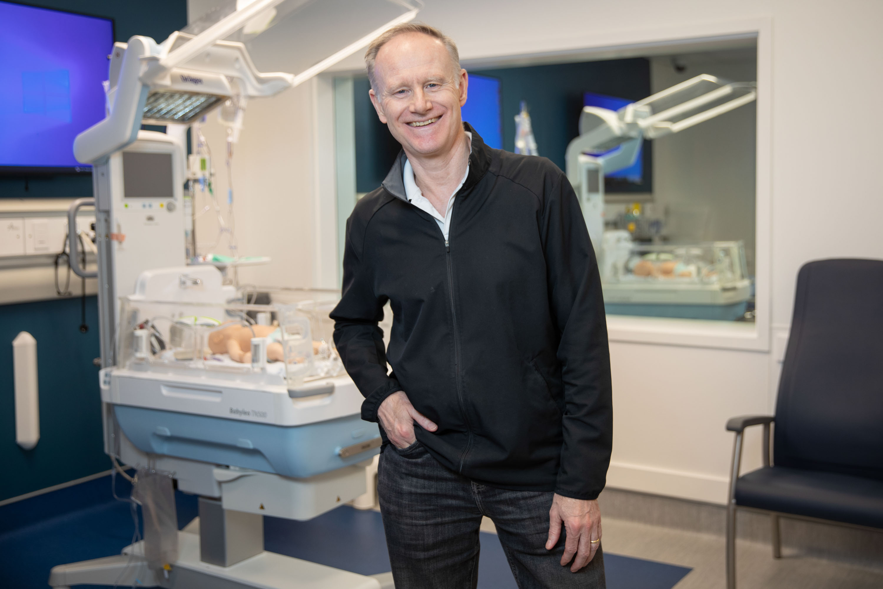 Dr. Jonathan Pirie stands in front of a hospital bed in the simulation centre, wearing a black jacket with one hand in his pocket.