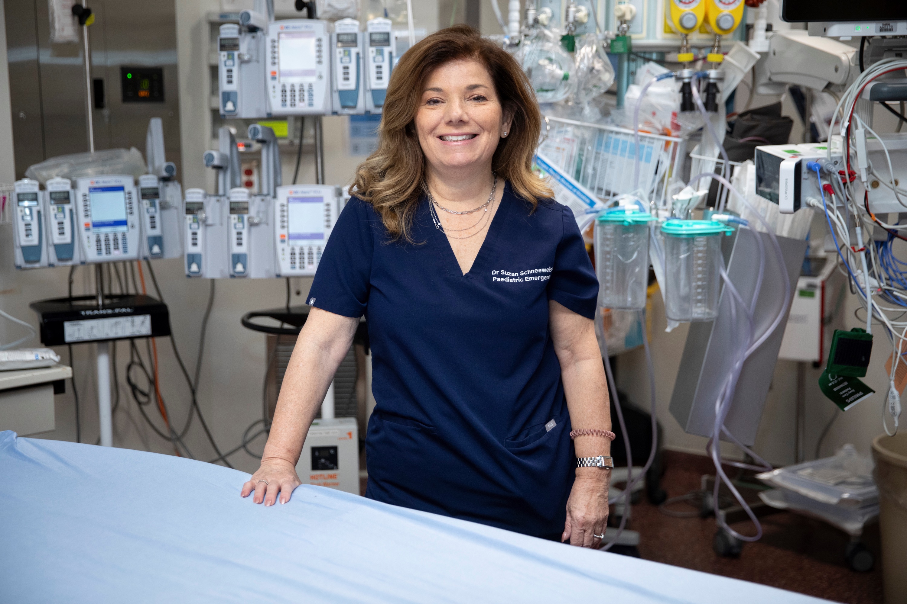 Dr. Suzan Schneeweiss, wearing navy blue scrubs, stands in front of a bed with a blue sheet on it in a trauma room in the emergency department. 