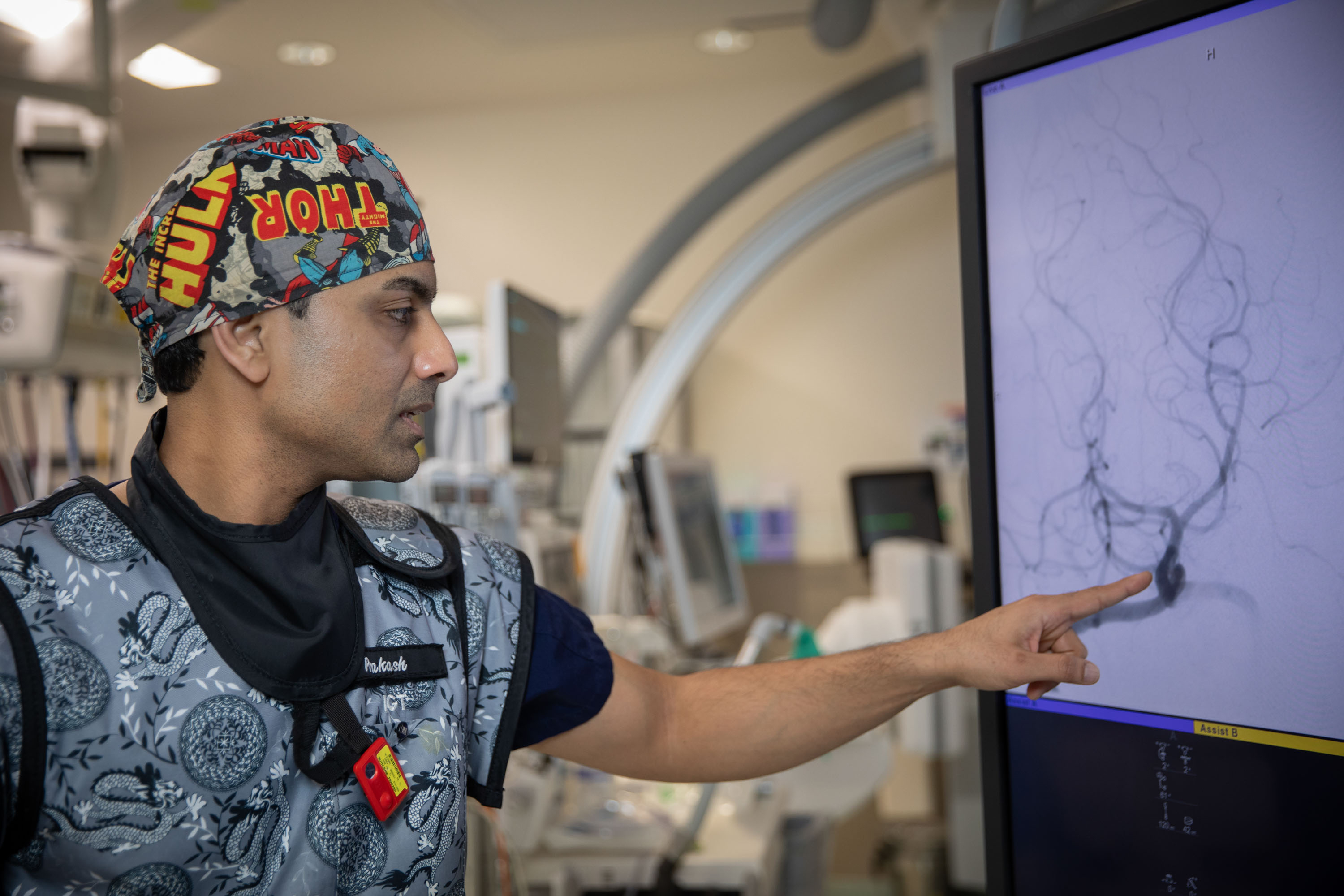 Interventional cardiologist in comic-themed surgical cap pointing to cerebral angiogram on monitor in catheterization laboratory.