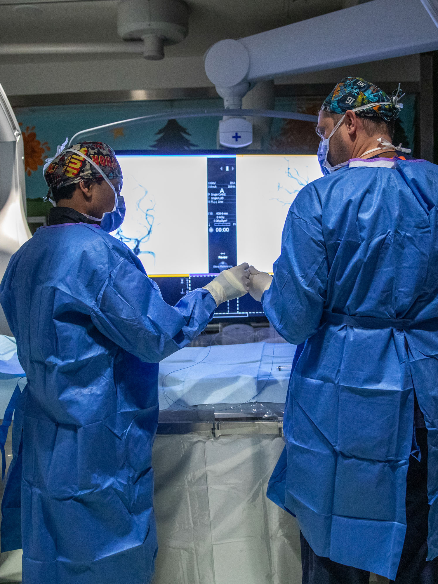Two surgeons in blue scrubs and patterned surgical caps perform a procedure using advanced medical imaging equipment in an operating room.