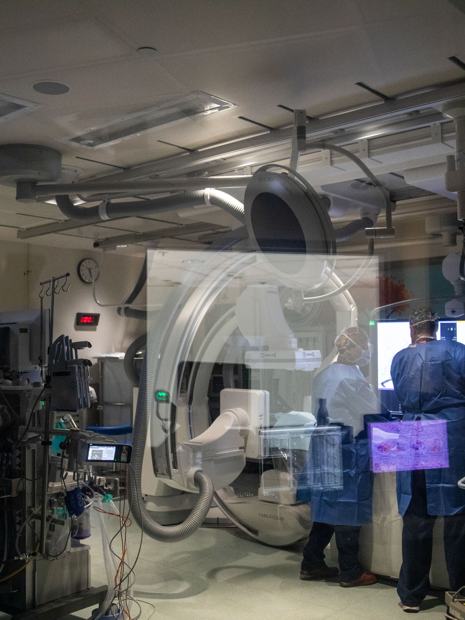Advanced cardiac catheterization laboratory with C-arm fluoroscopy equipment and medical staff performing interventional procedure with multiple monitoring screens.