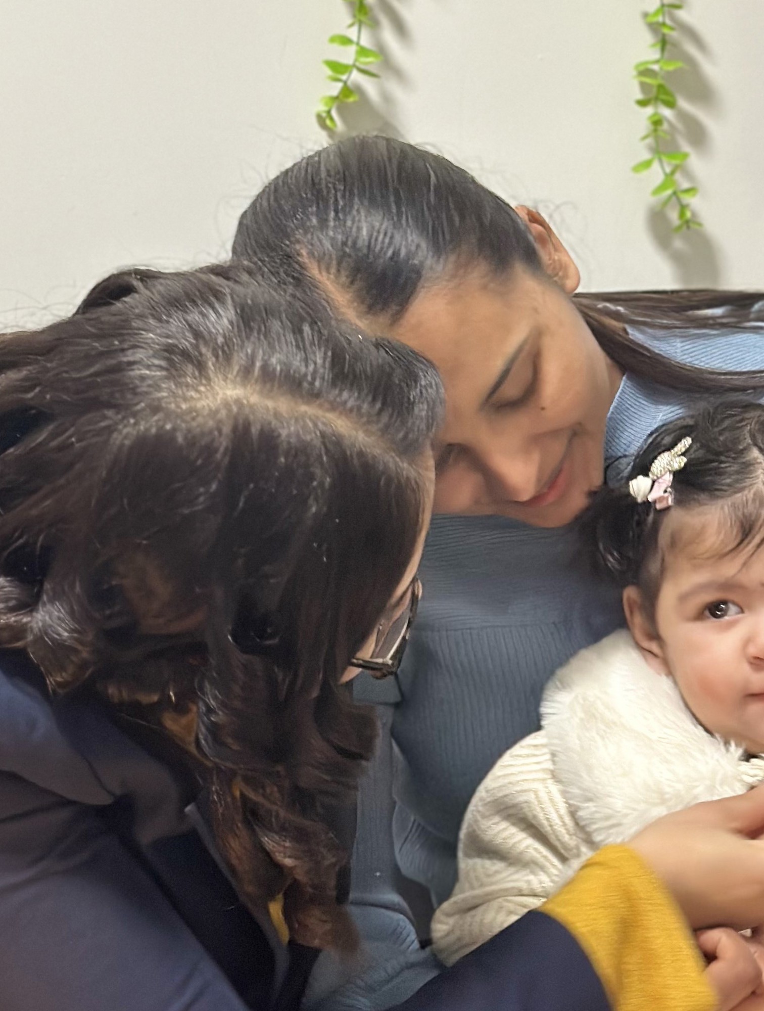 Two women holding and interacting with smiling baby in cream sweater with hair bows in comfortable home setting.