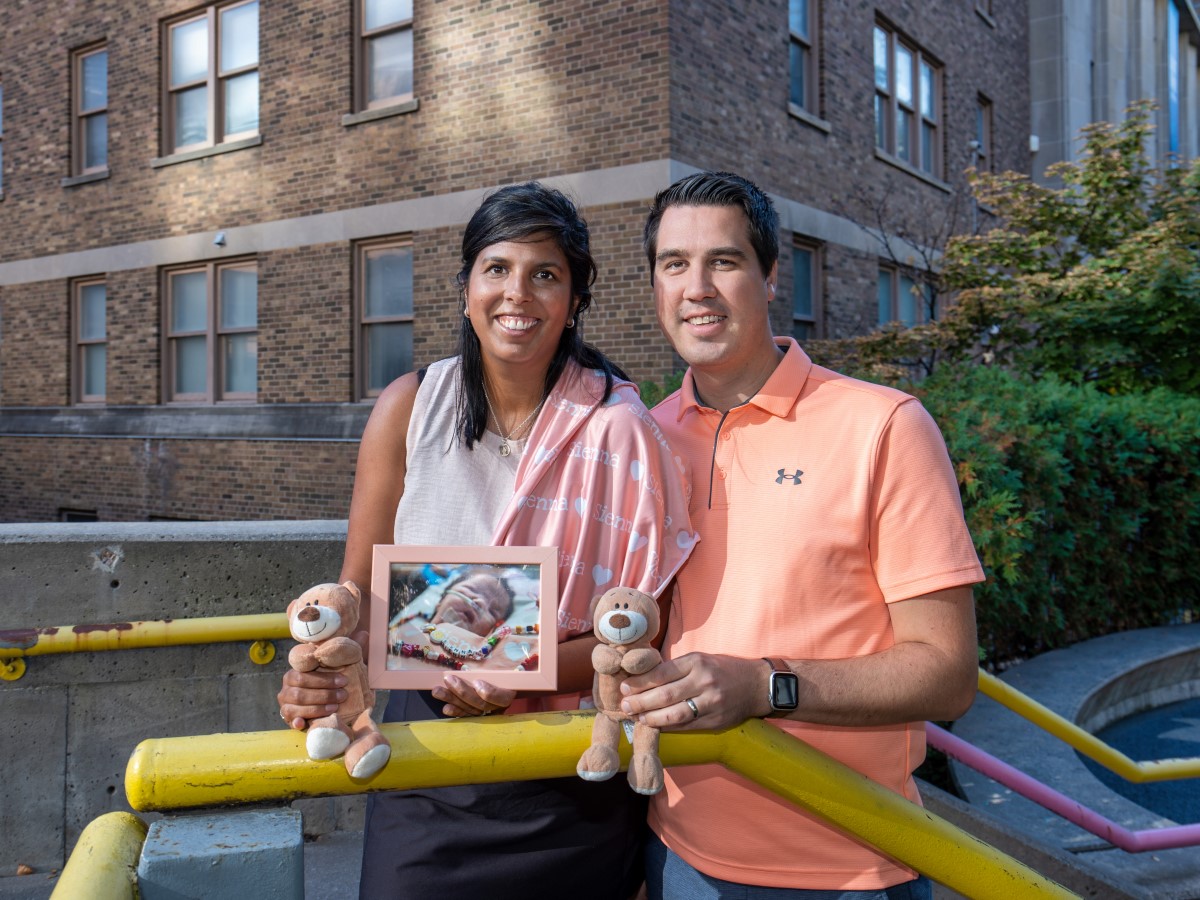 Brittany and Tyler, wearing peach, stand in front of a yellow railing. Brittany holds a framed photo of Sienna, and both hold a stuffed bear.