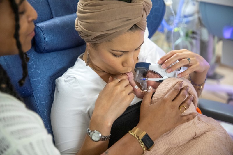 Person holding baby with breathing tube and medical equipment; another individual partially visible on the left.