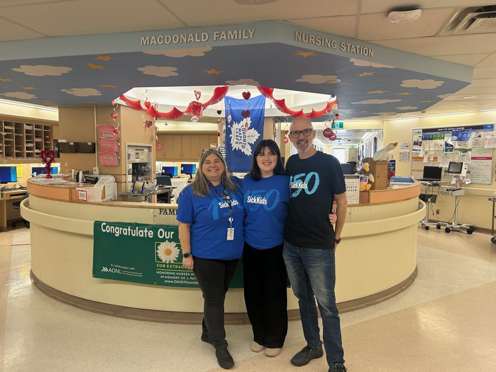 Kat, Liv and James wear SickKids 150 t-shirts, standing in front of the desk of SickKids' Cardiology unit.