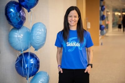 Krista Cardamone wearing a blue SickKids t-shirt standing in a hallway beside blue balloons.
