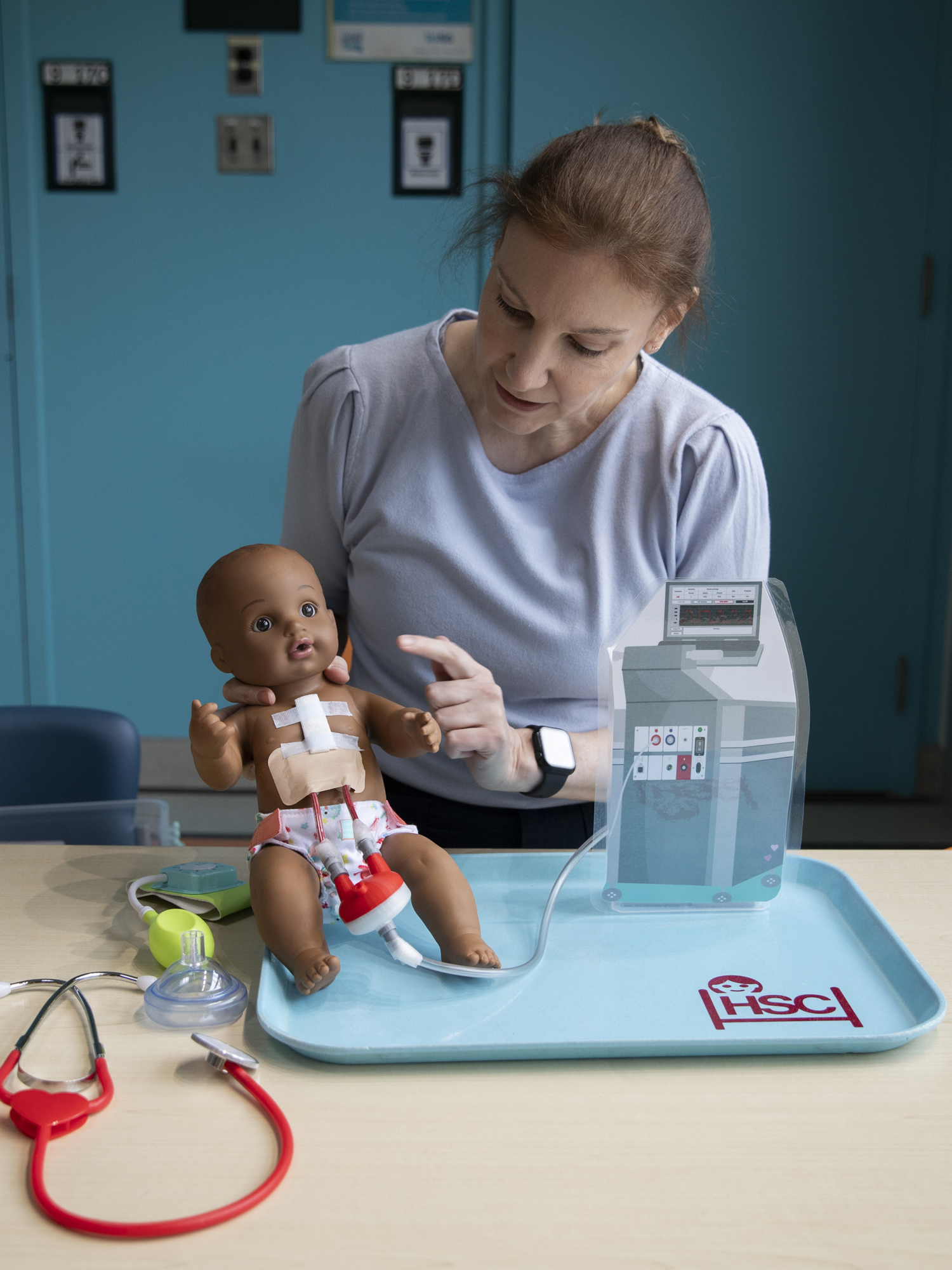 Natalie sits at a table and points at a doll that has a Ventricular Assist Device. There is a stethoscope and other medical equipment on the table.