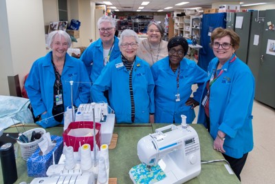Five women in blue jackets pose together at a sewing table.