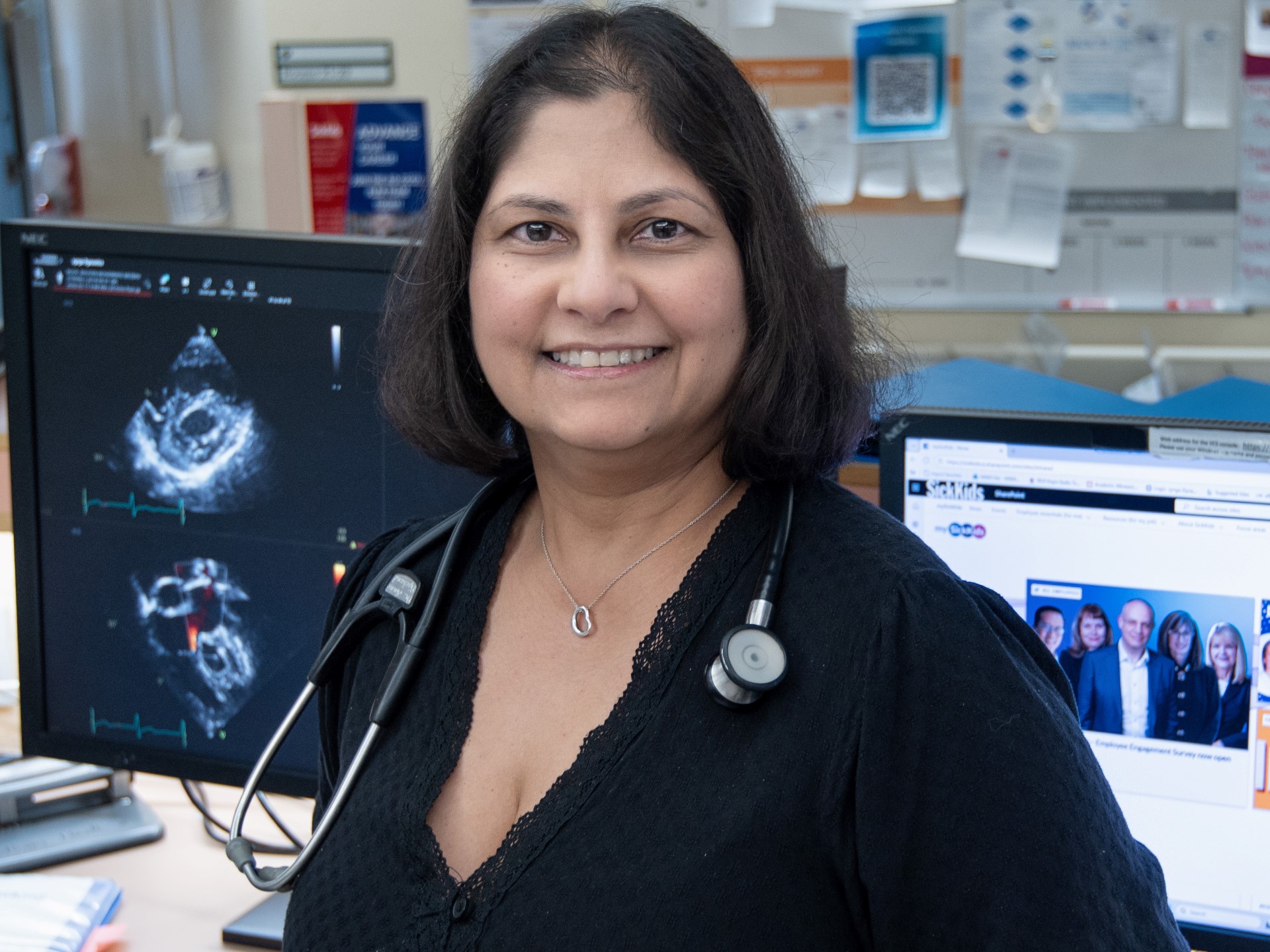 Wearing a black shirt and stethoscope, Dr. Anne Dipchand smiles in front of computer screens showing medical images. 
